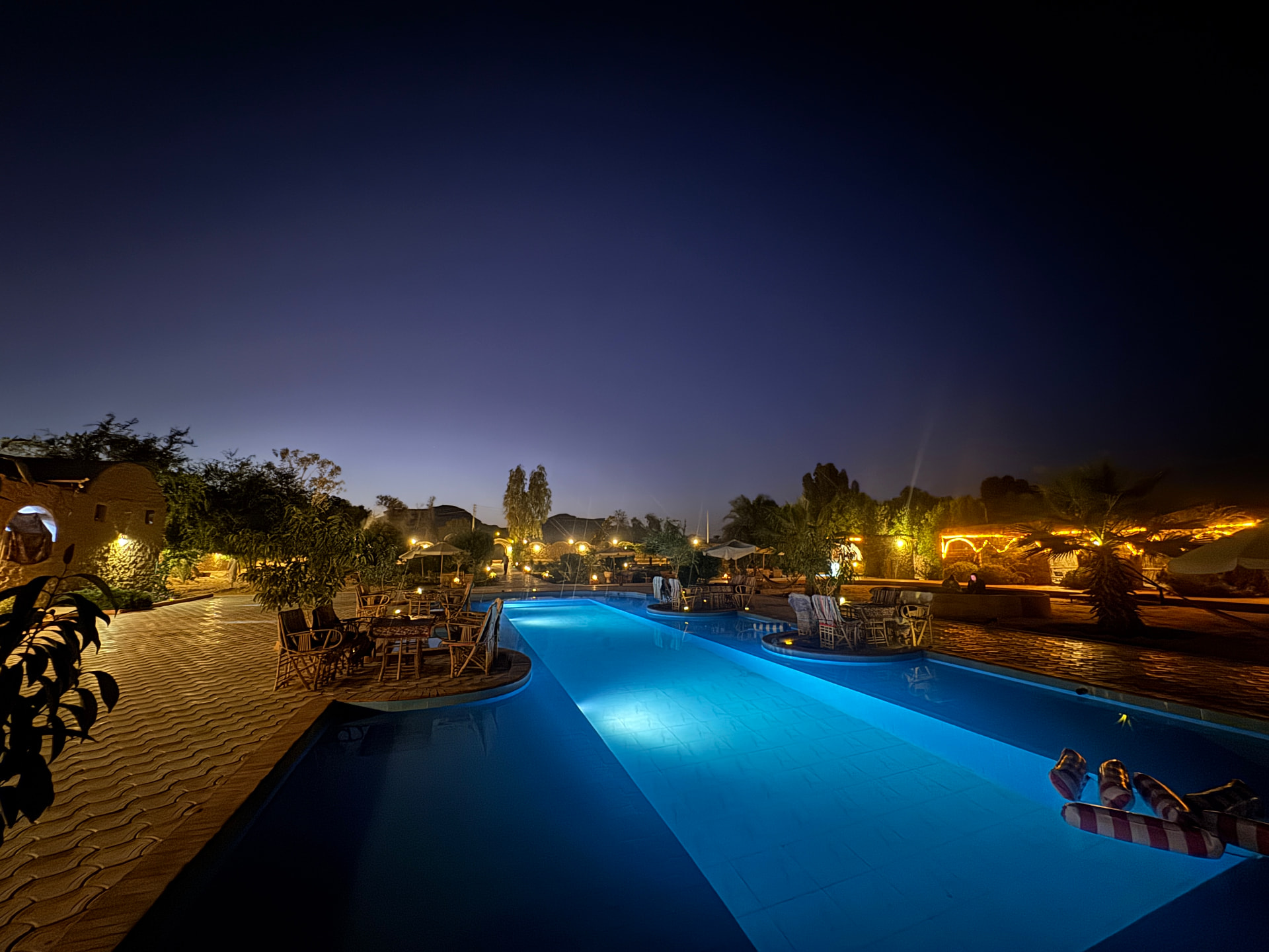 The Siwa pool glowing blue at night, lanterns in the gardens